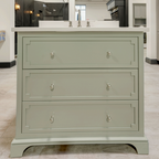 A three-drawer bathroom vanity cabinet in a muted sage green finish with silver metal knobs, shown with a white double-sink countertop in a showroom setting.