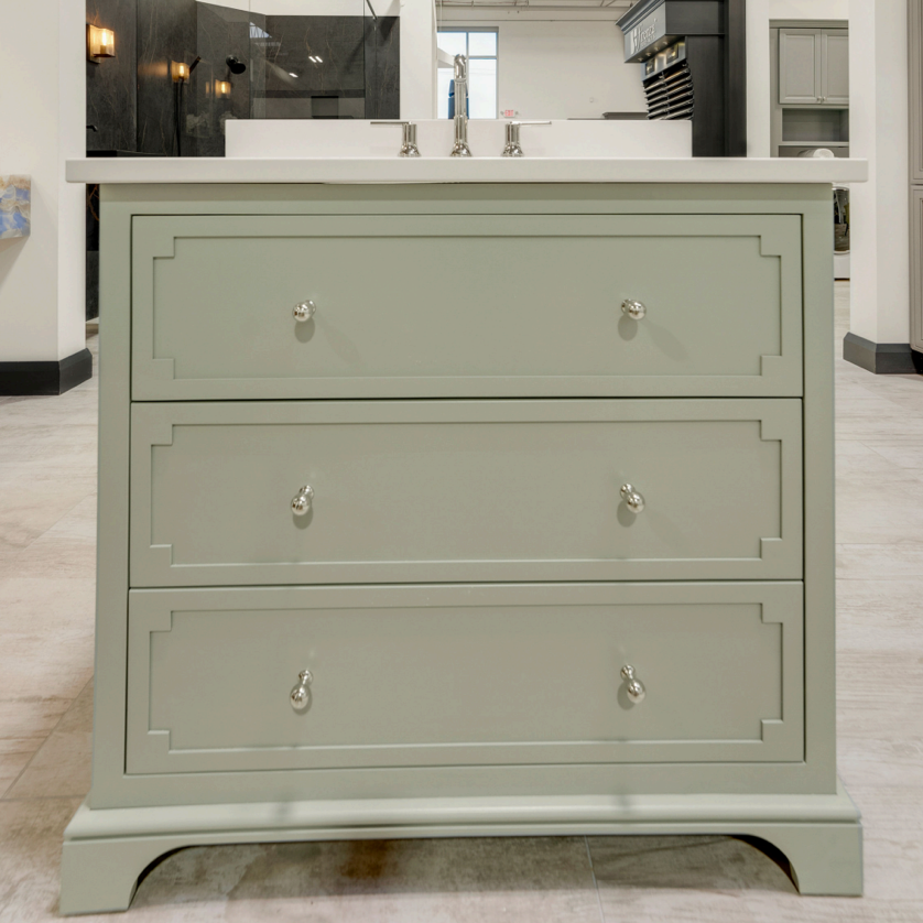 A three-drawer bathroom vanity cabinet in a muted sage green finish with silver metal knobs, shown with a white double-sink countertop in a showroom setting.