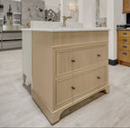 A light beige wood kitchen vanity cabinet with three drawers and small round metal knobs, topped with a white stone countertop and integrated sink, shown in a showroom setting.
