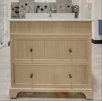 A light beige wood bathroom vanity cabinet with two drawers and dark metal knobs, set in a showroom with a white wall, marble tile backsplash, and the 'firenza' brand logo visible on the wall.