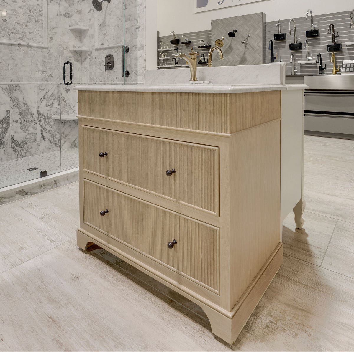 A light wood bathroom vanity with a white countertop and sink, shown in a showroom. The background features a marble shower enclosure and a wall sign reading 'firenza'.