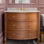 A classic wooden bathroom vanity with three drawers and brass knobs, set against a dusty mauve paneled wall with a matching mirror and gold faucet.
