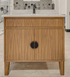 A modern bathroom vanity cabinet in warm wood with a white countertop and sink, shown in a showroom. The wall behind reads 'firenza' in blue.