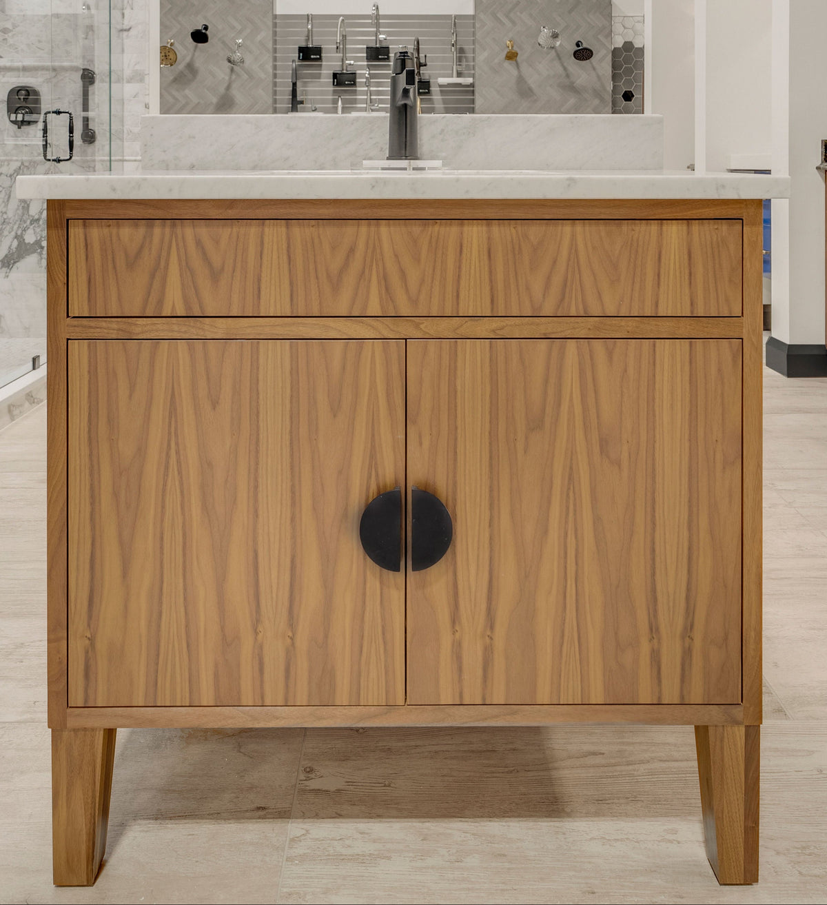A modern bathroom vanity cabinet in warm wood with a white countertop and sink, shown in a showroom. The wall behind reads 'firenza' in blue.